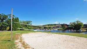 A sandy beach area with a volleyball net, a grassy field, and a lake with boats in the background.