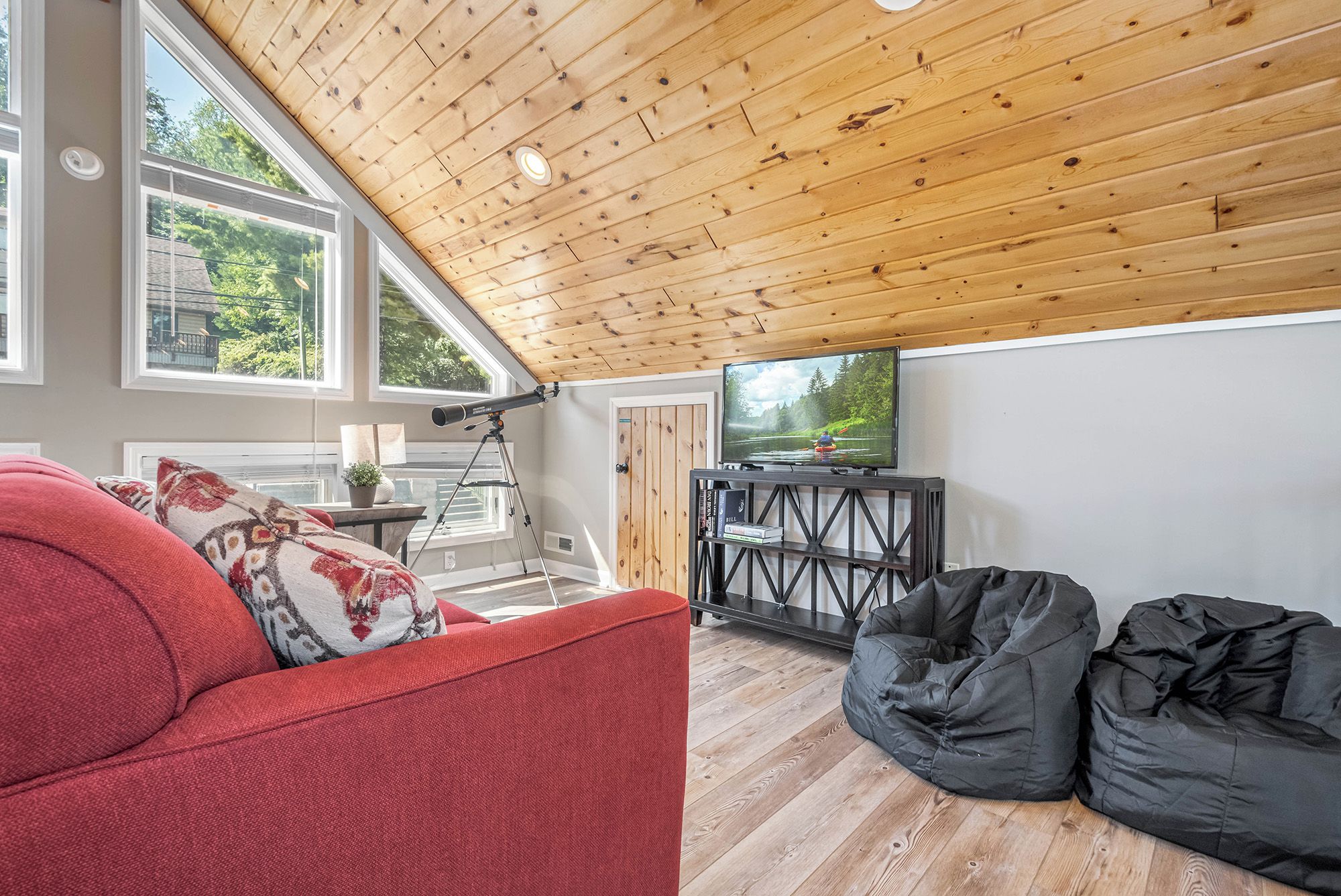A cozy living room with a wooden ceiling, a red sofa, black bean bags, and a television displaying a nature scene.