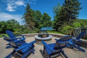 An outdoor seating area with Adirondack chairs around a stone fire pit, surrounded by lush greenery and a view of a lake. - Ski to Shore Chalet by Taylor Made Deep Creek (McHenry)