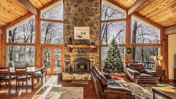 A cozy cabin interior with a stone fireplace, wooden furniture, and large windows offering views of the surrounding trees. - Slip Away Chalet by Taylor Made Deep Creek (McHenry)