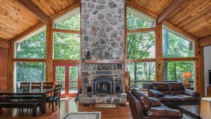 A spacious room with a stone fireplace, wooden ceiling, large windows, and a leather sofa. - Slip Away Chalet by Taylor Made Deep Creek (McHenry)