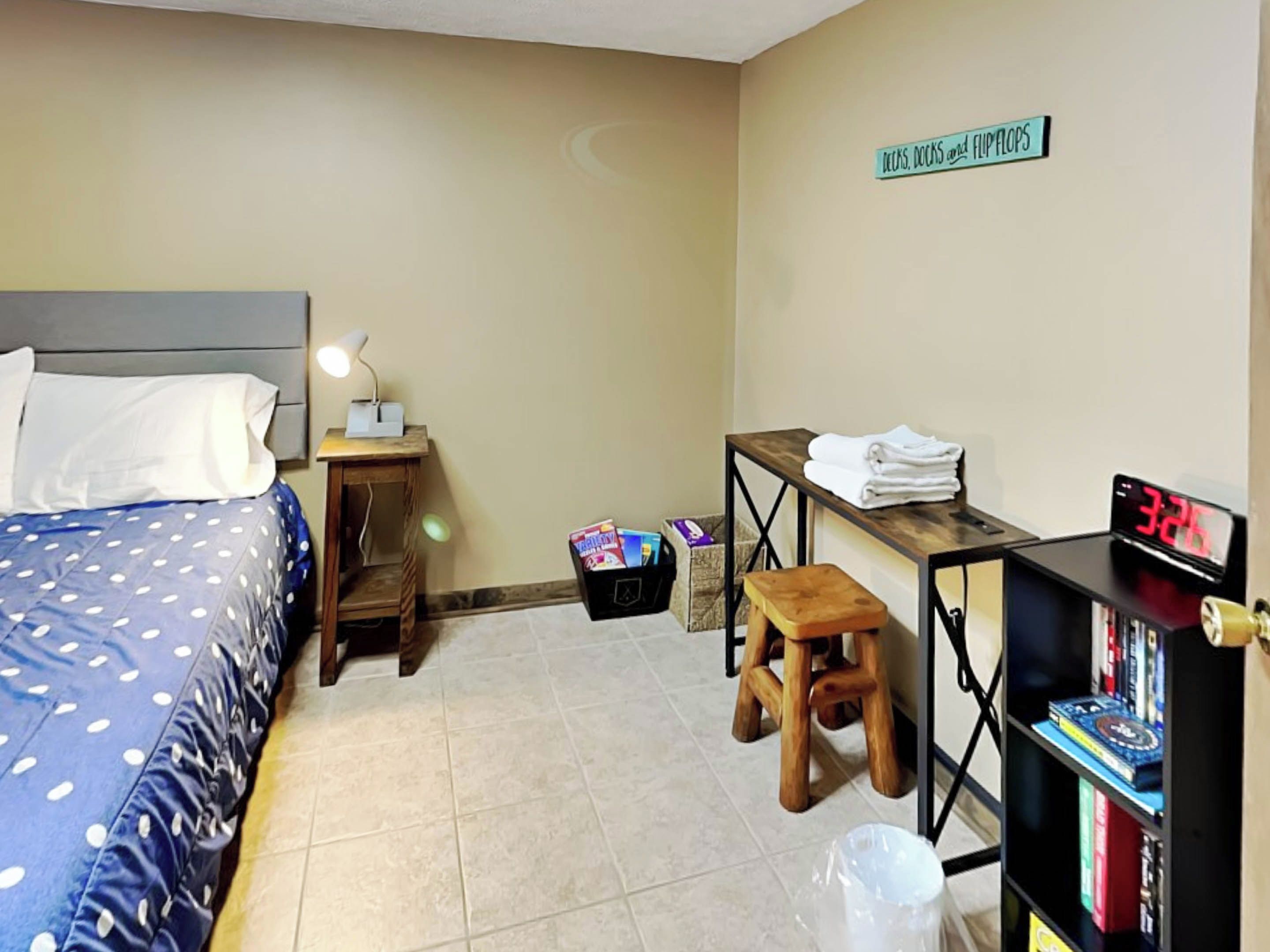 Quarto de hotel com uma cama, uma mesa, uma cadeira, uma estante e um letreiro na parede.