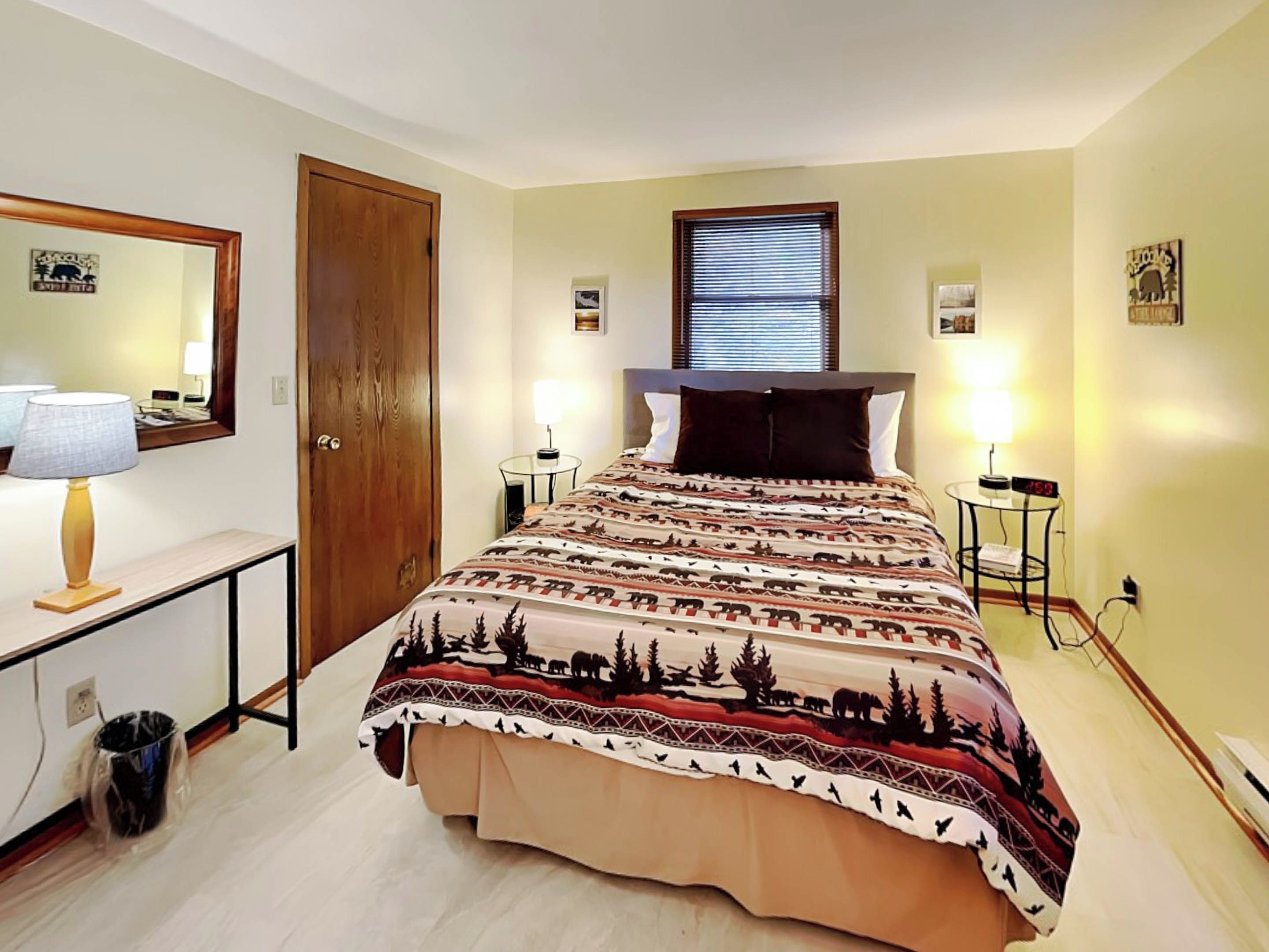 A bedroom with a bed, a wooden door, a bedside table with a lamp, a small table with a clock, and a window with blinds.