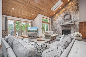 A spacious living room with a stone fireplace, large windows, and a television. - Solar Delight by Taylor Made Deep Creek (McHenry)