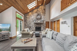 A spacious living room with a stone fireplace, wooden ceiling, and large windows offering a view of the outdoors.