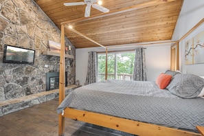 A bedroom with a stone fireplace, a bed with grey bedding, and a ceiling fan.