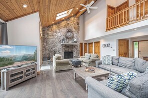 A spacious living room with a stone fireplace, wooden ceiling, and a balcony.