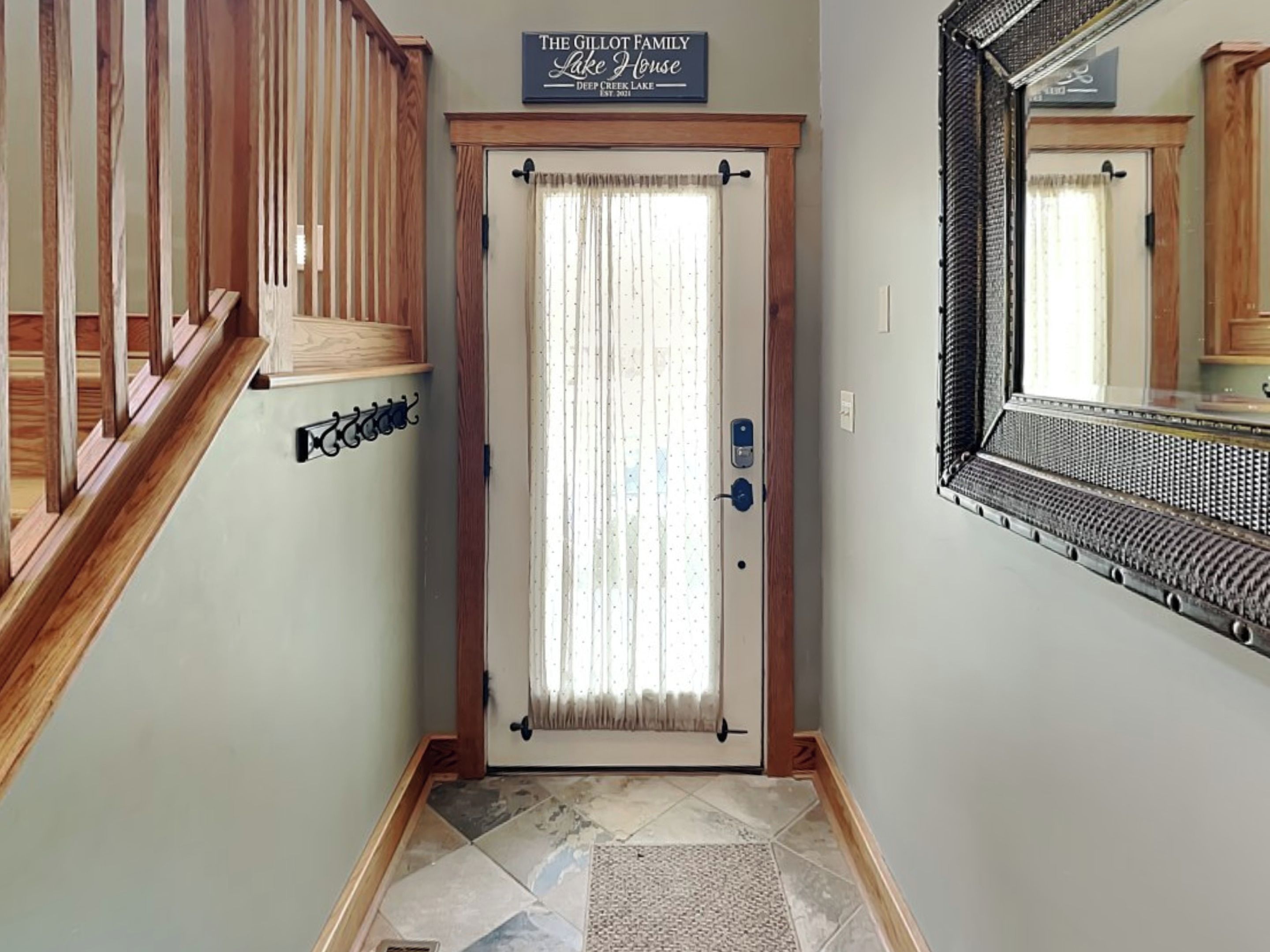 A hallway with a wooden staircase, a door with a sign reading "The Gillot Family Lake House Deep Creek Lake," and a mirror on the wall.