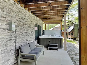 A covered outdoor area with a stone wall, a hot tub, a sofa, and a wooden deck. - Space Mountain by Taylor Made Deep Creek (McHenry)