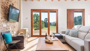 Amplia sala de estar con pared de piedra, vigas de madera, televisor de pantalla plana, sofá, mesa de centro y grandes puertas correderas de vidrio que dan a un jardín.