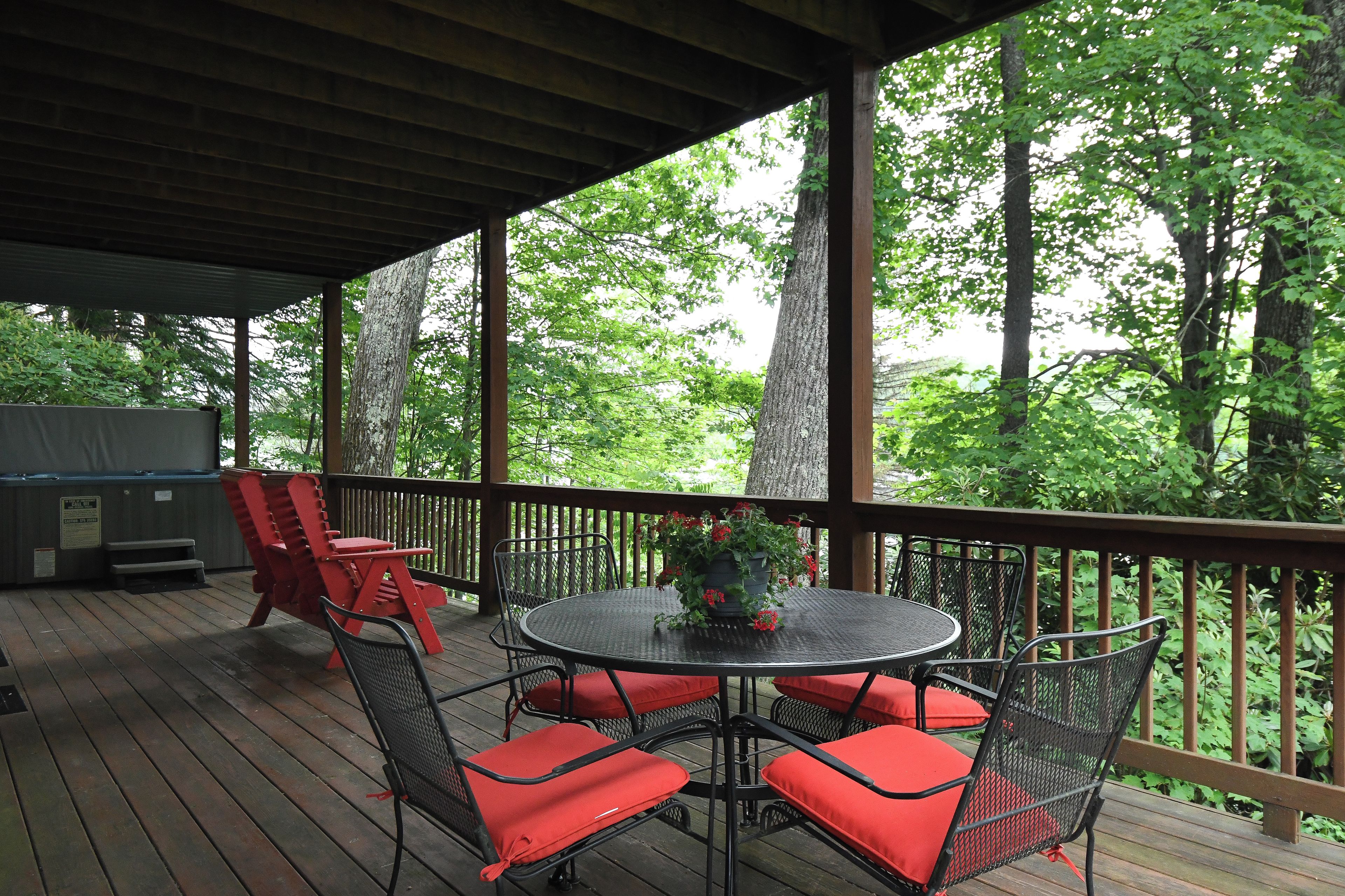 A covered outdoor deck with a round table and chairs, surrounded by a wooden railing and trees.