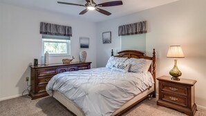 Quarto com cama, penteadeira, mesa de cabeceira, luminária, ventilador de teto e janela com cortinas.