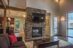 A living room with a stone fireplace, a flat-screen TV mounted above it, and a large window offering a view of the outdoors.