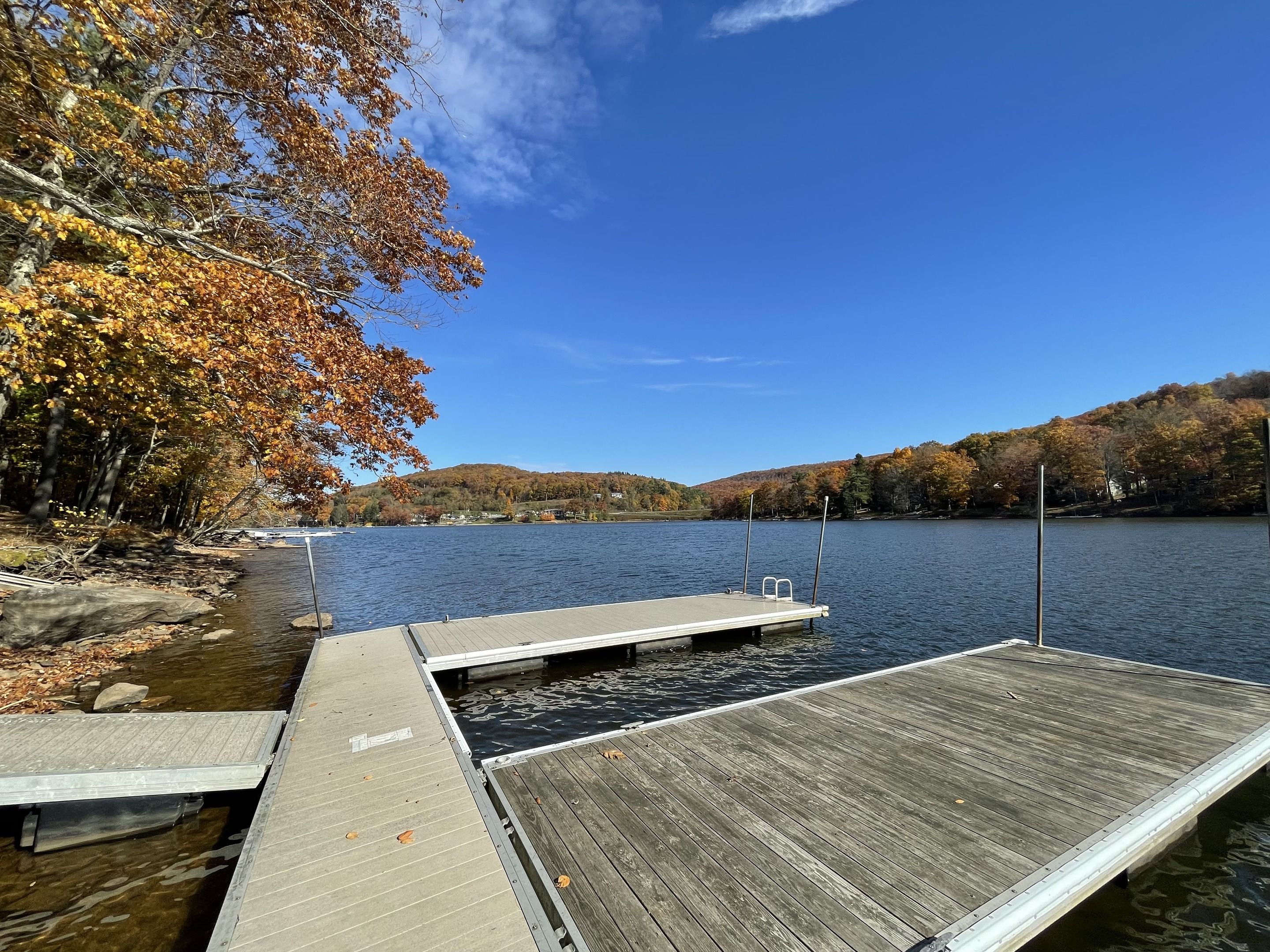 A wooden dock extending into a calm lake, surrounded by autumn-colored trees and hills in the background.