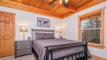 A bedroom with a wooden ceiling, a bed with a headboard, two bedside tables with lamps, a world map on the wall, and a window with a view of trees.