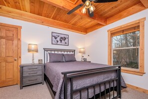 A bedroom with a wooden ceiling, a bed with a headboard, two bedside tables with lamps, a world map on the wall, and a window with a view of trees. - Stay Awhile by Taylor Made Deep Creek (McHenry)