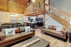 Amplia sala de estar con techo alto y vigas de madera, cocina con electrodomésticos modernos, y escalera con barandilla de madera.