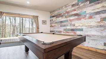A pool table in a room with a large window, a rustic wall, and a small clock.