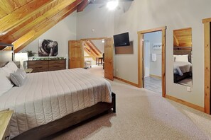 A bedroom with a large bed, wooden ceiling beams, a television, and a bathroom with a mirror. - Stay Awhile by Taylor Made Deep Creek (McHenry)