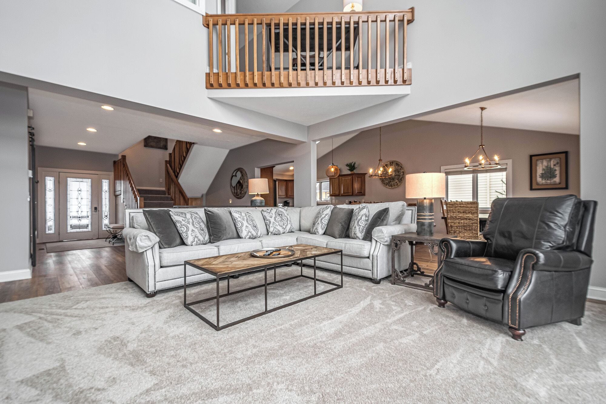 A spacious living room with a high ceiling, a wooden balcony, a large sectional sofa, a leather armchair, and a coffee table.