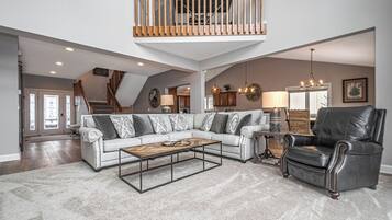 A spacious living room with a high ceiling, a wooden balcony, a large sectional sofa, a leather armchair, and a coffee table.