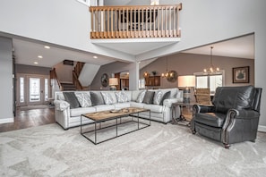 Ein geräumiges Wohnzimmer mit hoher Decke, einem Holzbalkon, einem großen Ecksofa, einem Ledersessel und einem Couchtisch.