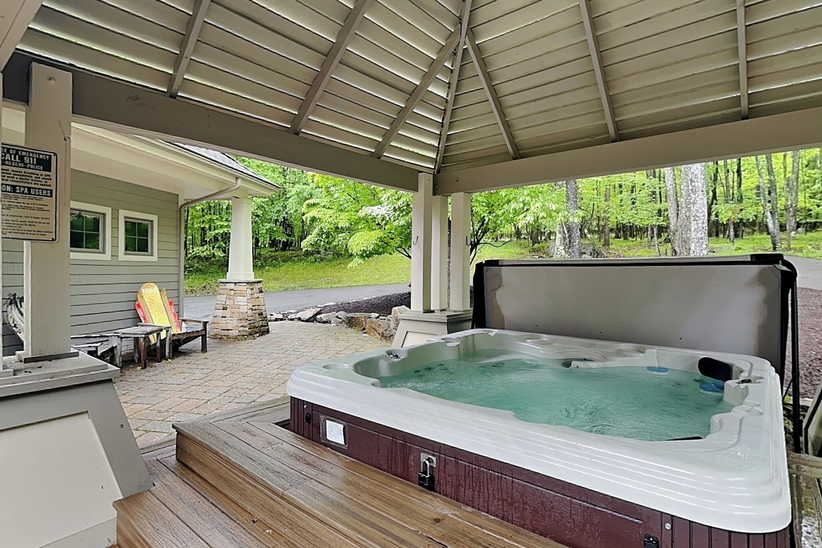 A covered outdoor area with a hot tub, wooden deck, and a house in the background.