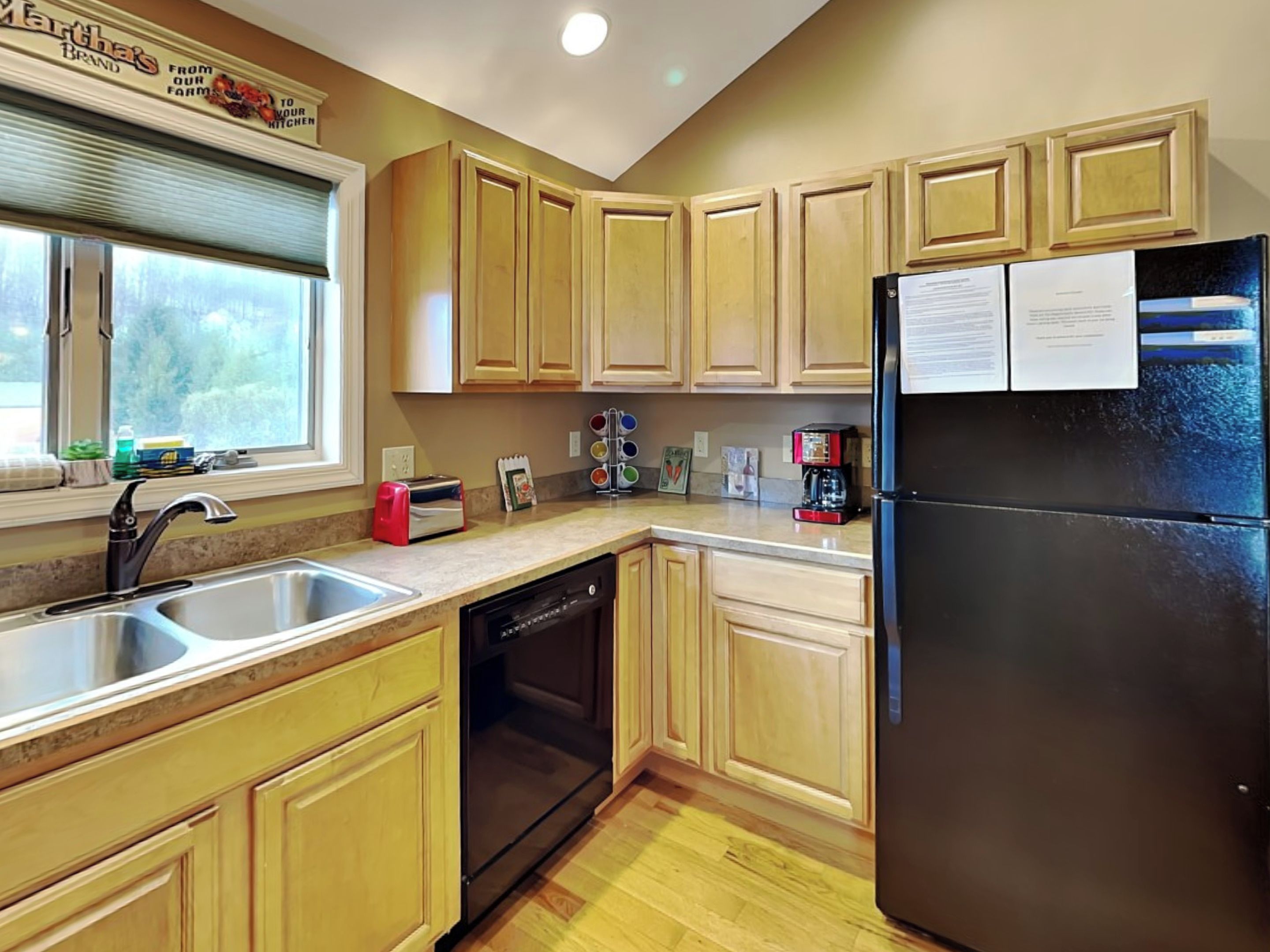 A kitchen with wooden cabinets, a black refrigerator, a dishwasher, and a window with a view of greenery.