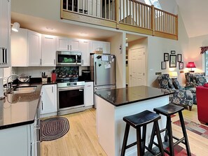 A modern kitchen with white cabinets, stainless steel appliances, a central island with stools, and a wooden staircase. - Summer Haven by Taylor Made Deep Creek (Oakland)
