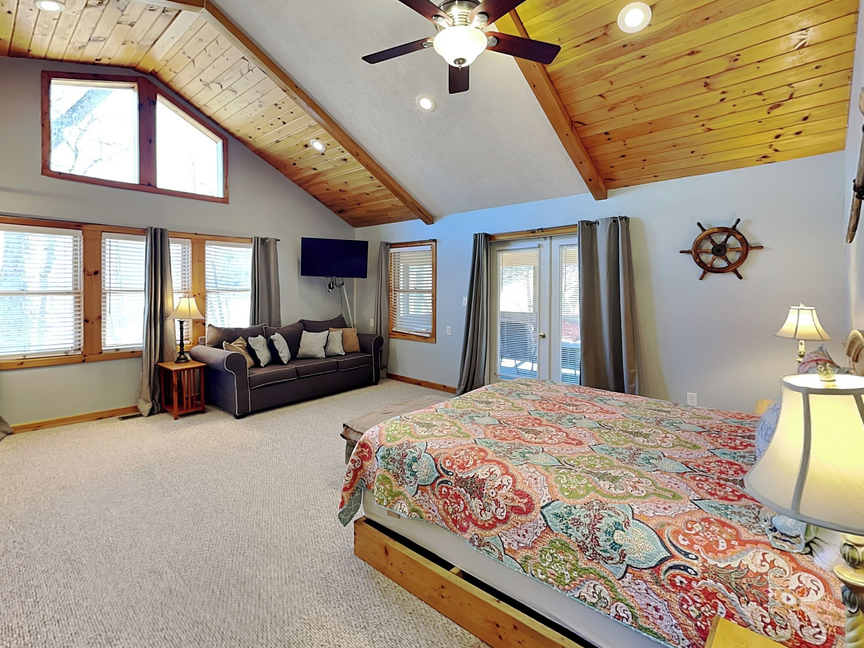 A bedroom with a bed, a sofa, a TV, a ceiling fan, and large windows.