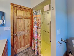 A bathroom with a wooden door, a shower with a colorful shower curtain, and a toilet with a towel rack. - Sunkissed Lodge by Taylor Made Deep Creek (McHenry)