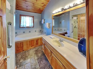A bathroom with a bathtub, a sink, wooden cabinets, a mirror, and a window with curtains. - Sunkissed Lodge by Taylor Made Deep Creek (McHenry)