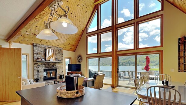 A spacious living area with a stone fireplace, large windows overlooking a lake, and a wooden ceiling. - Sunny Boy by Taylor Made Deep Creek (McHenry)