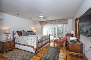 Um quarto com cama, ventoinha no teto, janela com cortinas, mesa de cabeceira, cadeira e cômoda.