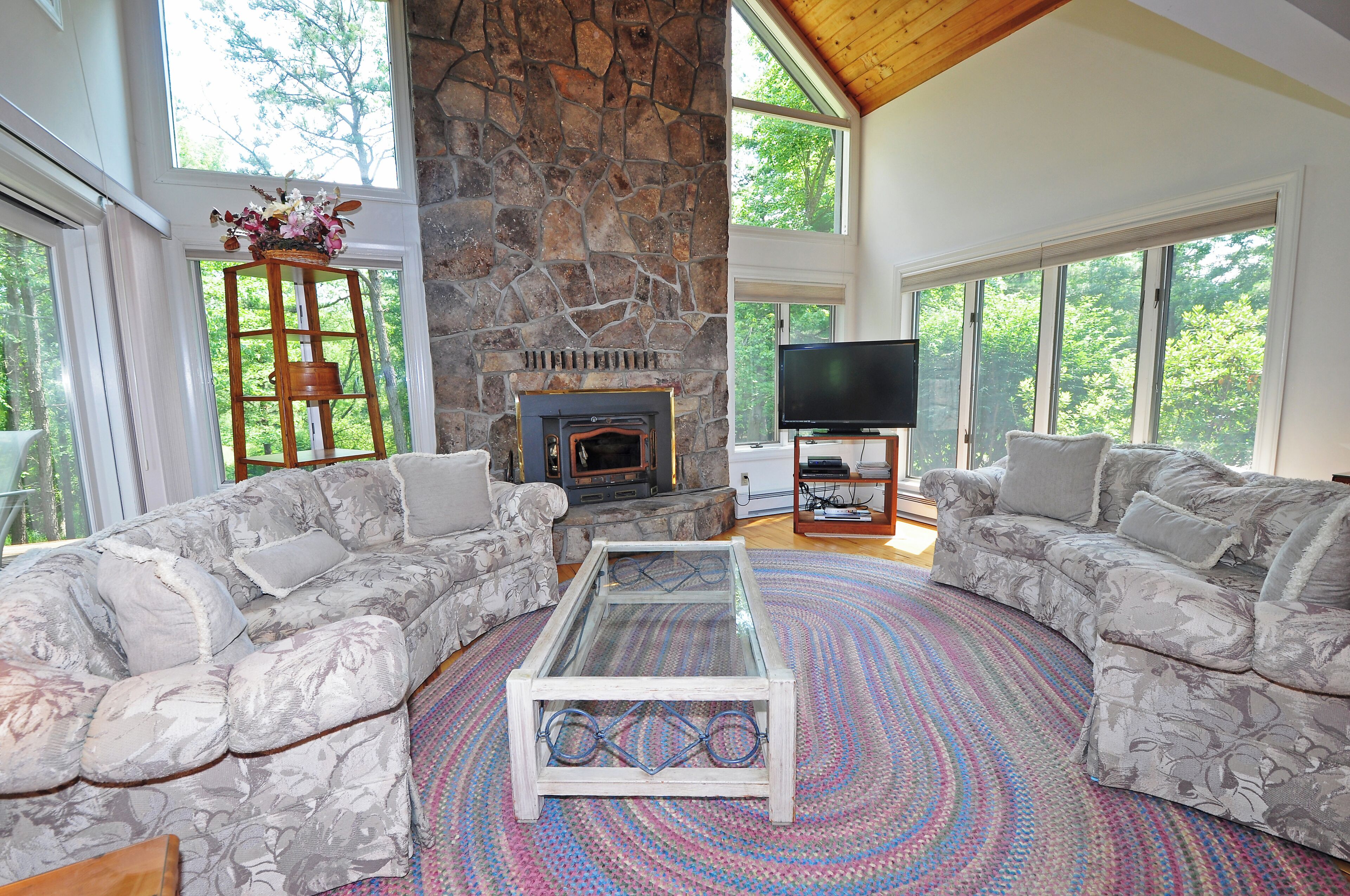A spacious living room with a stone fireplace, two grey patterned sofas, a glass coffee table, and a flat-screen TV.