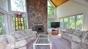 A spacious living room with a stone fireplace, two grey patterned sofas, a glass coffee table, and a flat-screen TV. - Sunrise by Taylor Made Deep Creek (Oakland)