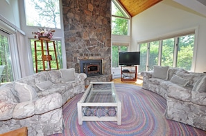 Un salón amplio con chimenea de piedra, dos sofás grises con estampado, una mesa de centro de vidrio y un televisor de pantalla plana.