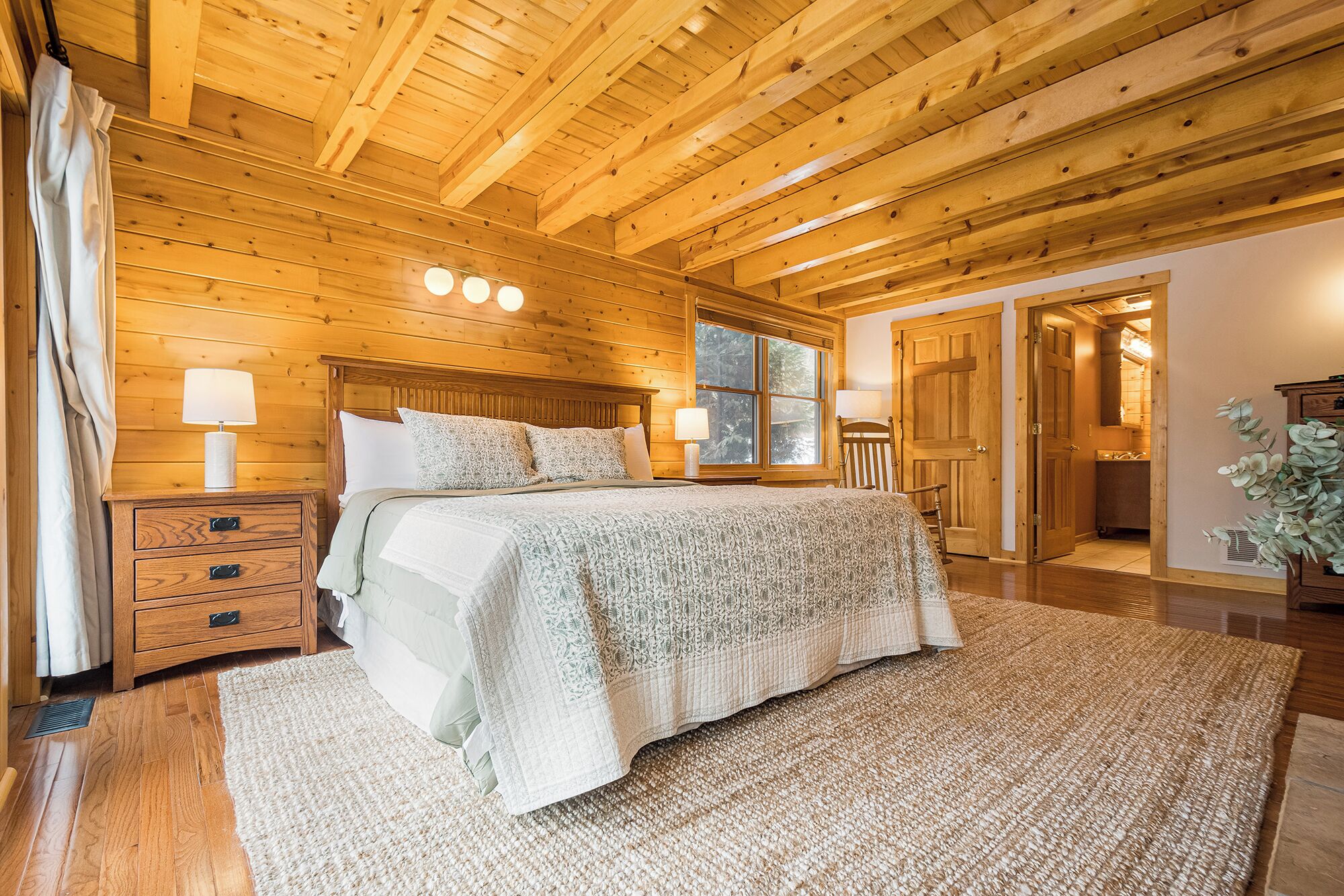 A wooden cabin bedroom with a large bed, two bedside tables, a window, and a door leading to a bathroom.