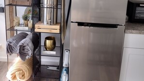 Et kompakt køkken med et rustfrit stål køleskab, en mikrobølgeovn og en lille vask. Der er en metalreol med diverse køkkenartikler og en kurv oven på mikrobølgeovnen.