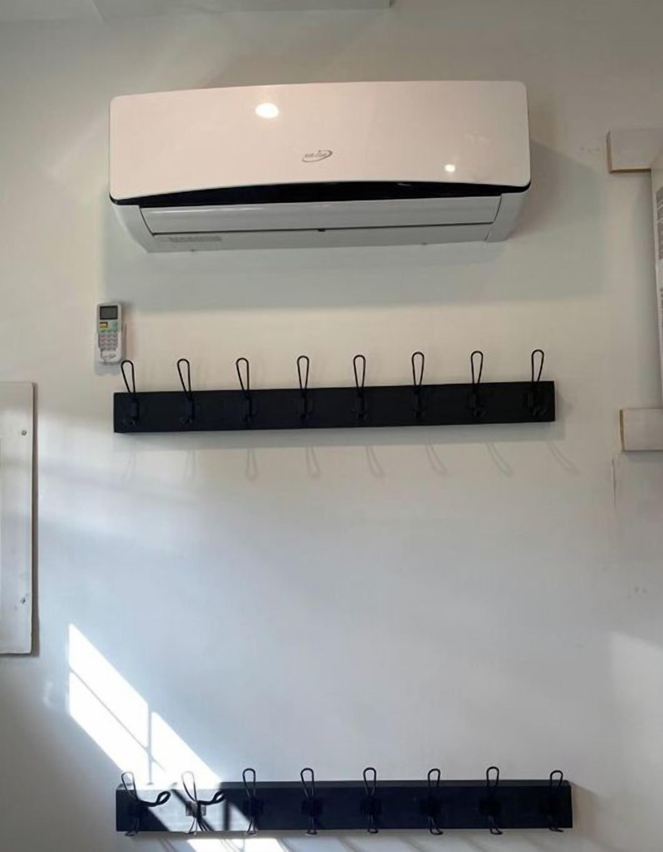 A white air conditioner mounted on a wall, a black shelf with hooks below it, and a whiteboard to the left.