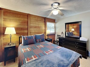 A bedroom with a wooden wall panel, a ceiling fan, a bed with a patterned quilt, a nightstand with a lamp, a dresser with a mirror, and a window with blinds. - Swan's Nest by Taylor Made Deep Creek (Swanton)