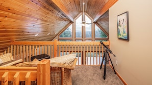 A cozy attic bedroom with wooden walls, a sloped ceiling, a ceiling fan, a large window, a telescope, and a framed painting.