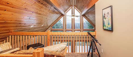 A cozy attic bedroom with wooden walls, a sloped ceiling, a ceiling fan, a large window, a telescope, and a framed painting.