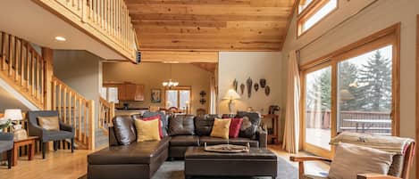 A spacious living area with a wooden staircase, a sectional sofa, and a large window offering a view of the outdoors.