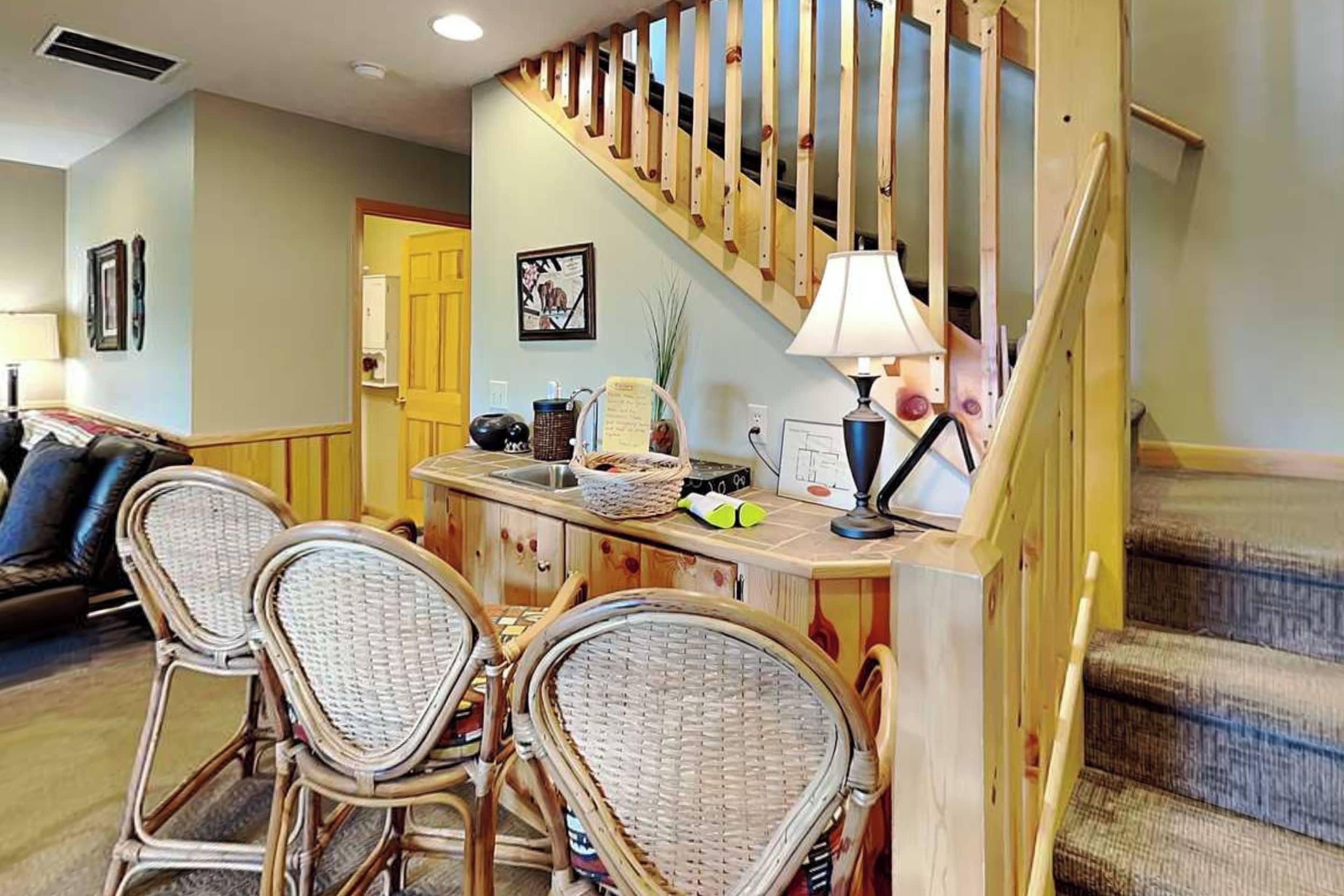 A living room with a wooden staircase, wicker chairs, a dining table with a lamp, and a sofa.