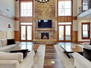 A spacious living room with a stone fireplace, large windows, and a high ceiling. - The Haven by Taylor Made Deep Creek (Oakland)