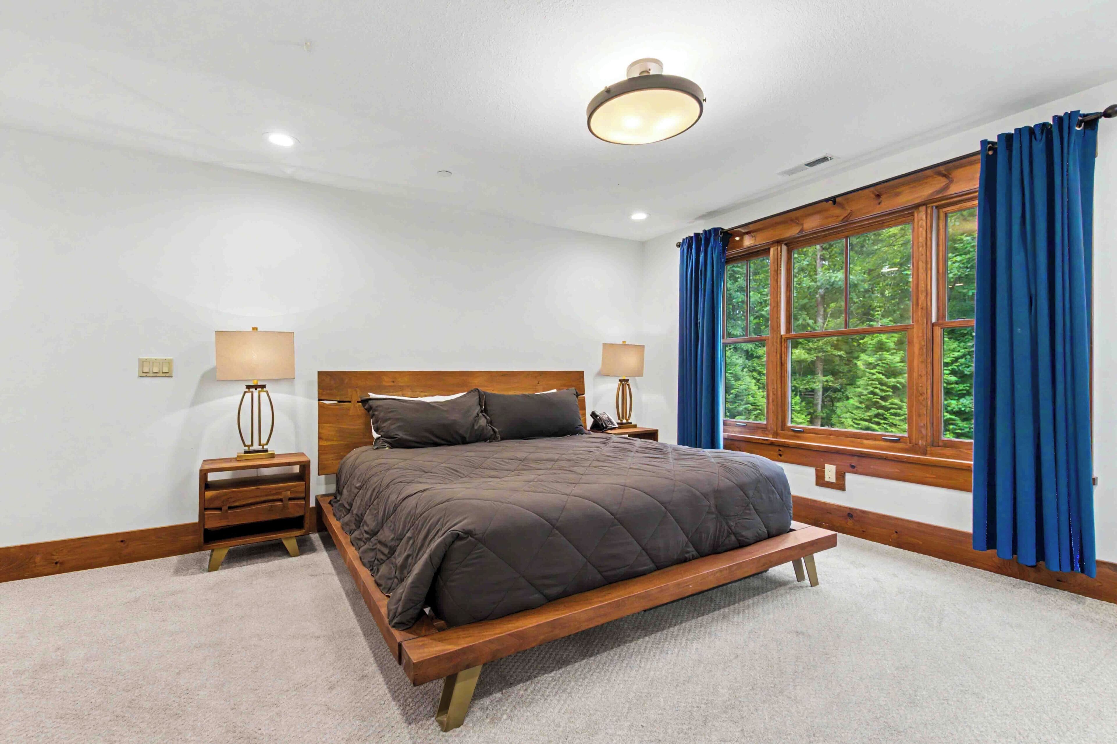 A bedroom with a wooden bed, two bedside tables with lamps, a window with curtains, and a ceiling light.