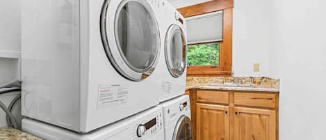 A laundry room with stacked front-loading washers and dryers, wooden cabinets, a granite countertop, and a window with a view of greenery.