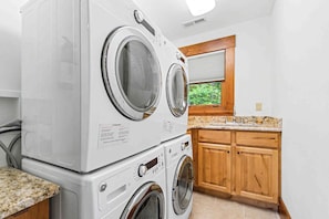 A laundry room with stacked front-loading washers and dryers, wooden cabinets, a granite countertop, and a window with a view of greenery. - The Haven by Taylor Made Deep Creek (Oakland)