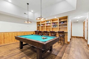 A pool table with chairs, a bookshelf, and pendant lighting in a room with wooden flooring and white walls. - The Haven by Taylor Made Deep Creek (Oakland)
